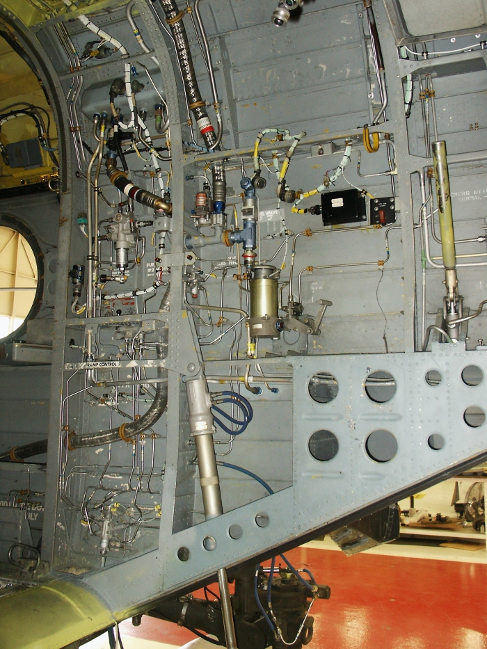 The right inside ramp area of CH-47D Chinook helicopter 85-24336 showing the number one and two flight boost hydraulic system and utility hydralic sytem fill module, the manual hand pump used to pressure the utility hydraulic system, the chip fuzz burn off module, the power steering module, and the number two engine cross feed and engine fuel shutoff valves. The long silver cylinder, center and bottom, is the right side ramp actuator.