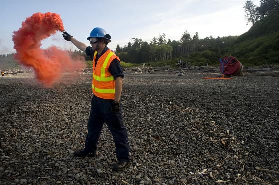 Standing near the buoy, Seaman Kevin Tkoch, District 13 Aids to Navigation Hardware, pops a smoke flare to help the CH-47 Chinook helicopter crew judge wind speed and direction before touching down on the beach in La Push, Washington.